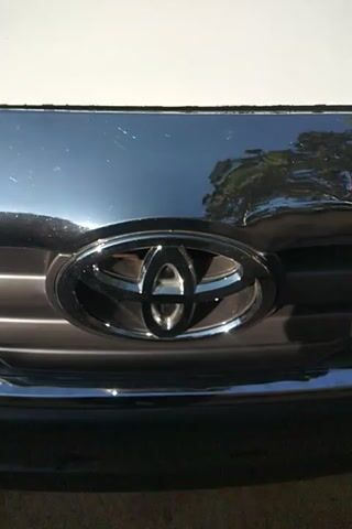 The video shows a close-up of a white Toyota vehicle. The camera first focuses on the front grill with the Toyota emblem in chrome. It then pans to the left, showing a portion of the windshield with a small decorative object affixed to the inside of the glass and a windshield wiper. The background appears to be an outdoor setting with trees or buildings reflected in the vehicle's shiny surfaces. The lighting is bright, suggesting daytime.