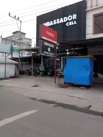 The video shows a street scene with several buildings and vehicles. A black minivan drives past on the left. A woman on a white scooter is visible in the middle of the road. Several other motorcycles and cars are also present. Buildings on the right include one with a large sign that reads "AMBASSADOR CELL". There are also signs for "NUBIA" and "QATAR COPY". The sky is overcast.