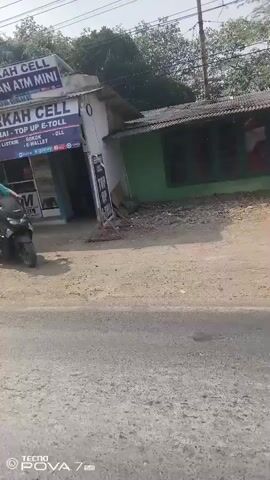 The video shows a person riding a motorcycle through a street with shops and houses. One shop, "Berkah Cell," sells mobile phone services and ATM mini services. There is a red and white gas pump visible. The street is unpaved in some sections and paved in others. Cars and a truck are also seen on the road. The video is shot from the perspective of someone riding a motorcycle.