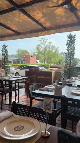 The video shows an empty outdoor dining area. The tables are neatly set with plates, glasses, silverware, and napkins. A QR code is visible on one table. The chairs are made of dark wicker. In the background, through an open-air awning, there is a street scene with cars, trees, and a building with a sign that says "Bocconcino." The sky is blue and sunny. A ceiling fan rotates above the tables. The camera pans slightly to the right. A person's arm and hair appear briefly on the right side of the frame.