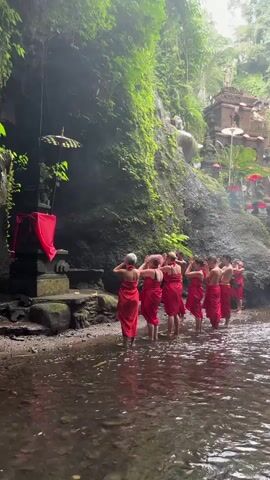 A group of eight individuals, all wearing vibrant red sarongs, stand facing a moss-covered cliffside with a small stream in the foreground. Their backs are to the camera, and they appear to be performing a ritual, with most of them touching their foreheads or hair. The cliff is adorned with small, ornate structures, possibly shrines, and there are several decorative umbrellas. A large statue, partially obscured by foliage, is visible on the cliff. The environment suggests a lush, natural setting, possibly a temple or sacred site in a jungle or mountainous region. The lighting is dim, indicating a shaded area, possibly due to heavy tree cover or a cave-like overhang.