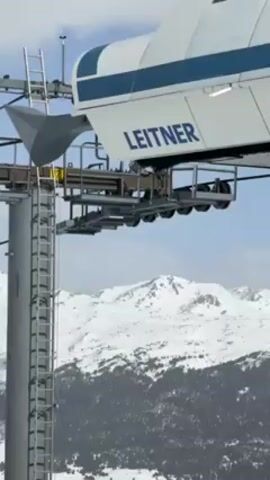 The video shows a ski lift tower with a "LEITNER" sign on top. There is a ladder attached to the tower. Skiers ride the lift, with snowy mountains visible in the background.