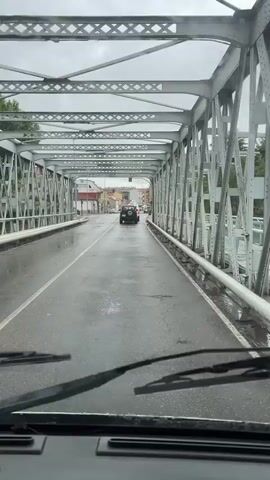 The video shows a point-of-view shot from a vehicle driving across a bridge. The bridge is made of metal girders and has a wet asphalt road surface, indicating it may be raining or has recently rained. There is a single vehicle, a dark-colored SUV, ahead of the camera vehicle. The SUV has a spare tire mounted on its rear. The road has white lane markings. On either side of the bridge, there are railings and views of buildings and trees. Traffic lights are visible in the distance. The sky appears overcast.