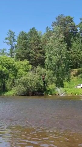 A video captures a person in a black outfit performing a backflip into a body of water. The person is seen in mid-air, executing the flip, before splashing into the water. The environment is lush and green, with trees and foliage lining the riverbank. The water is clear and rippling, with the sky above appearing blue and partly cloudy. The person is alone in the water.
