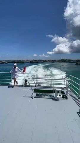 The video shows the wake of a boat from the perspective of the stern deck. A woman wearing a white outfit and hat stands to the left, leaning against the railing. In the background, a coastline with buildings and other boats is visible. The sky is blue with scattered clouds. The deck is grey and has white railings. The water is turquoise, churning into white foam from the boat's movement. A man wearing a white shirt walks into the frame from the right side of the screen.