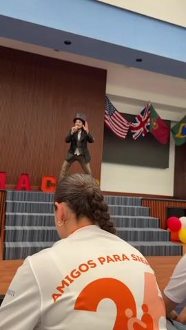 The video shows a young male performer on a stage, dressed in a dark coat, vest, hat, and khaki-colored pants. He holds a microphone and appears to be speaking or performing for an audience. The stage is decorated with large, block letters that spell out "MAC." Behind the stage is a wooden-paneled wall and several flags, including the flags of the United States, the United Kingdom, Argentina, and Spain.

The camera is positioned from the perspective of someone in the audience, focusing on the back of a person wearing a white shirt with "AMIGOS PARA SIEMPRE" printed on it in orange lettering. A small boy is also visible to the left of the person. The room has wooden floors and the person behind the camera appears to be seated on the floor.