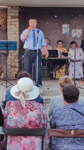 A man in a light blue checkered shirt, dark blue tie, and dark pants stands on a raised platform, speaking into a microphone. He is holding a second microphone in his left hand. To his left, another man in a suit and hat stands near a music stand. Behind the main speaker, a table is set up with a laptop and two women are seated, one wearing a floral dress and the other a white patterned dress. The audience, mostly women wearing hats, is seated in chairs facing the stage. The background appears to be an outdoor setting with a brick wall and some draped fabric.