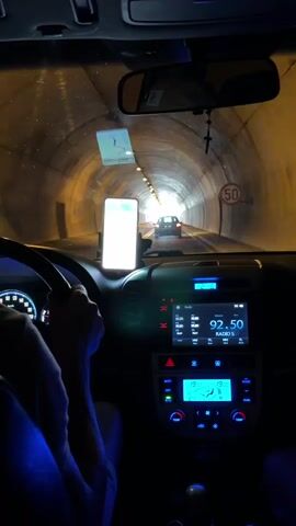The video shows the interior of a car driving through a tunnel. A person's hands are visible on the steering wheel. A smartphone is mounted on the dashboard displaying a navigation app. The car's radio is on, and the climate control system is visible. Another car is ahead in the tunnel. The tunnel walls are illuminated by overhead lights. A speed limit sign indicating 50 is visible on the right side of the tunnel.