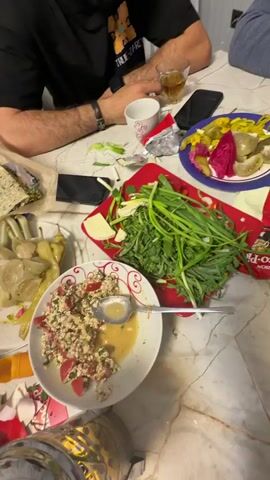 The video shows a group of people sitting around a table playing backgammon while enjoying a meal. The table is covered with various dishes of food, including salads, herbs, and bread. There are also drinks and a teapot on the table. A man is actively playing backgammon, moving the pieces on the board. Other people are either eating, talking, or watching the game. The room is well-lit and appears to be a dining area in a home.