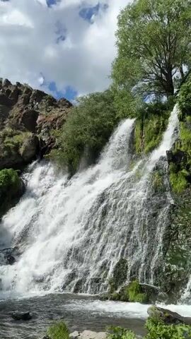 This is a vertical video of a multi-tiered waterfall cascading down a rocky, moss-covered cliff. Lush green trees are visible at the top of the waterfall and surrounding the area, with a clear blue sky and scattered white clouds overhead. The water is white and frothy as it plunges into a pool at the bottom, with some rocks visible in the foreground. The video shows the water flowing and splashing, creating a dynamic and natural scene.
