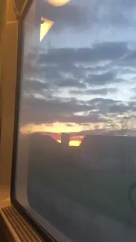 The video shows a view from a train window at sunset. The sky is a mix of blue and gray clouds with a bright orange sun low on the horizon. The sun's light illuminates the landscape below, casting silhouettes of trees and buildings. The train is moving, as evidenced by the passing scenery. The inside of the train car is visible as the frame of the window. The overall scene is peaceful and serene.