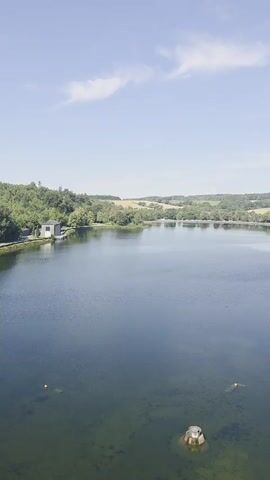 A drone view shows a wide, calm lake on a clear day. In the foreground, the water is clear enough to see submerged objects. In the middle distance, there are two orange buoys. On the left side of the lake, a small, white building sits near the shore. The shoreline is lined with trees. In the background, rolling hills covered in greenery and a small village with houses and buildings are visible under a bright blue sky with scattered white clouds. The drone slowly pans to the right, revealing more of the tree-lined shore and the village.