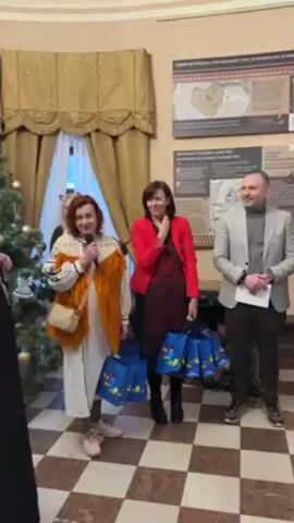 Three people stand in a room. To the left, a woman with red curly hair and a microphone stands in front of a Christmas tree, wearing an embroidered dress with an orange vest and tan purse. In the middle, a woman in a red blazer over a black patterned dress holds several blue gift bags. To the right, a man in a gray suit holds a white paper. The room has a black and white checkered floor. On the wall are informational panels in English and another language. The woman with the microphone appears to be speaking. The woman in the red blazer smiles and touches her neck. The man in the gray suit smiles.