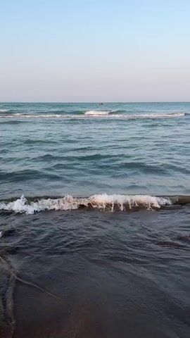 The video captures a low-angle view of ocean waves rolling onto a dark sand beach under a clear, pale blue sky. The water is a deep blue-green, with white foam cresting the incoming waves. In the distance, a small object, possibly a person on a jet ski or a distant boat, is visible on the water. The tide is coming in, with waves washing over the sand and creating ripples. The lighting suggests it might be early morning or late afternoon, as the sky is soft and the sun is not intensely bright.
