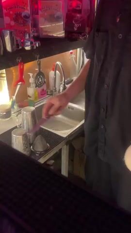 A person wearing a black collared shirt is preparing a drink behind a bar. They are holding a coconut and using a tool to pierce the top of it. In the background, there is a sink and various bar equipment.