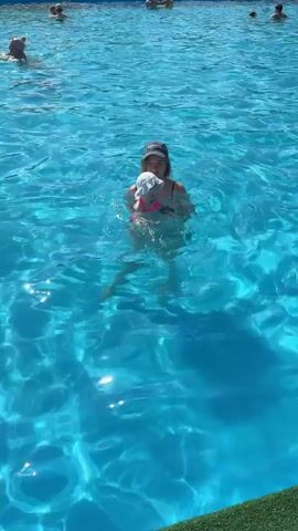 A woman in a black swimsuit and a baseball cap is holding a baby in a swimming pool. The baby is wearing a white hat and a pink swimsuit. The woman is holding the baby with her right arm and supporting its back with her left hand. The baby appears to be smiling and looking towards the woman. They are in clear blue water. There are other people in the background, also in the pool. The lighting suggests it is daytime and sunny.