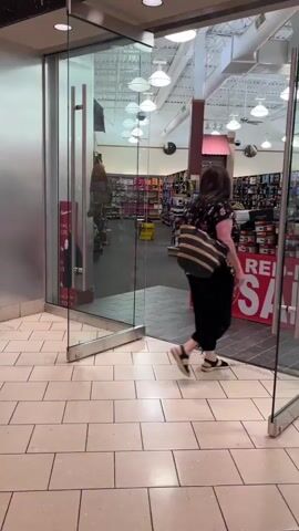 A person is exiting a shoe store in a mall. The store has a large glass door with a metal handle. Inside, there are shelves stocked with shoes and a sign that reads "50% OFF". The mall hallway has tiled flooring and a silver pillar. Two people are walking away from the camera down the hallway. The hallway is mostly empty with some store windows visible. The lighting is fluorescent and appears to be daytime.