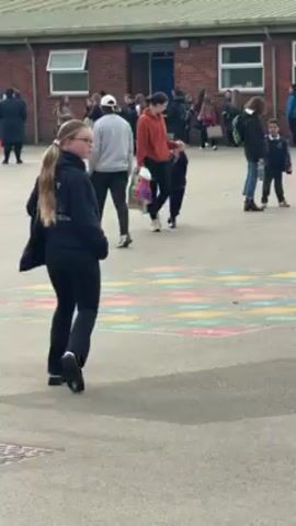 The scene is an outdoor schoolyard with a brick building in the background featuring windows and a blue door. A group of people, mostly children and some adults, are scattered around the paved area. Some are walking, while others are standing and talking. In the foreground, a young girl with long blonde hair in a ponytail, wearing a black hooded sweatshirt and black pants, walks away from the camera with a black bag slung over her shoulder. On the ground near her is a colorful hopscotch grid painted on the pavement.  A drain cover is visible in the bottom left corner of the frame. The weather appears to be overcast.