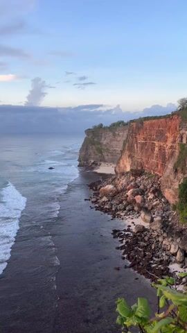 The video presents a scenic view of a coastline with tall, reddish-brown cliffs meeting the ocean. The sea stretches out under a sky of light blue with some white clouds, fading to a darker blue-gray horizon. White-capped waves roll towards the shore, crashing against the rocky base of the cliffs. The base of the cliffs is a mix of sandy areas and large boulders. Vegetation is seen atop the cliffs. No people are visible in the scene.