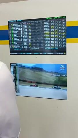 The video shows a close-up of two screens in what appears to be a racing control room. The top screen displays a live timing and scoring feed for a GT World Challenge GT3 race, showing driver names, lap times, gaps, and other race data. The bottom screen shows a live video feed of the race itself, with two GT3 cars on track. One car is red and the other is white and blue. The camera is focused on the screens, with a person's shoulder visible in the lower left corner of the frame. The room has white walls with yellow and blue stripes.