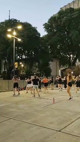 A dance fitness class of around 15 people of various ages is in progress in an outdoor plaza at dusk. Most participants wear workout attire, typically shorts and athletic tops, and sneakers.  The group follows the instructor, who is visible in the center, performing dance steps.  The plaza is paved with large concrete squares and surrounded by trees and old buildings.  Streetlights illuminate the area.  Some people are observing the class from nearby benches. A pink water bottle is visible on the ground next to the group.  The sky is dusky.  The overall impression is an urban, outdoor exercise session.