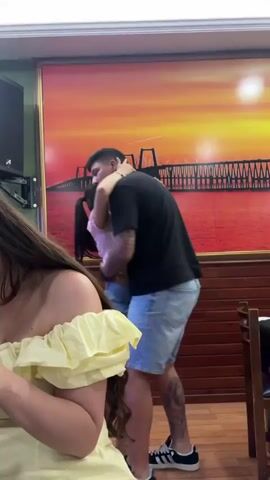 The video shows a young couple embracing. The man is wearing a black t-shirt and blue shorts, and the woman is wearing a pink crop top and denim shorts. They are in what appears to be an indoor setting, with a large framed picture of a bridge at sunset on the wall behind them. To the left, a woman with long brown hair is partially visible, wearing a yellow off-the-shoulder top. The lighting suggests it may be daytime, possibly late afternoon or evening, given the sunset image. The overall atmosphere seems casual and intimate.