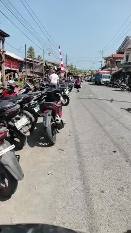 A person wearing a white shirt and dark shorts is standing near a parked motorcycle in a street lined with shops and parked motorcycles. A red and white barrier is visible in the distance. The ground is paved. The sky is clear and blue.