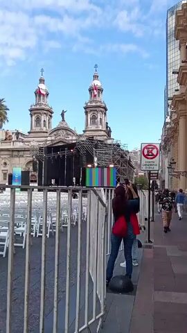 The video shows a scene of people taking photos in front of a large church building. The church has two prominent towers with Chilean flags on top. A stage is set up in front of the church with a large screen displaying color bars. A barrier separates the sidewalk from an area filled with white folding chairs. Several people are walking on the sidewalk, and one woman is taking a photo of the stage with her phone. A sign is visible on the side of the sidewalk with a red circle and a diagonal line through it, indicating a restriction of some kind. The sky is blue with some clouds, and there are modern buildings in the background.