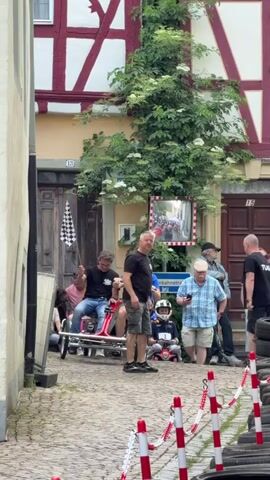 A video captures a street scene with a historical building featuring a brown and white facade and red wooden beams. In front of the building, a large green vine with white flowers climbs the wall. Several people are visible in the video, including two men in the foreground who are preparing to race in a tandem bicycle-like vehicle. One man in a black shirt holds a checkered flag, while the other man in a blue shirt is seated on the vehicle. Other individuals are standing around, some watching, and some appearing to be part of a team, wearing shirts with "TEAM" printed on them. A small child in a helmet sits on a toy vehicle in the background. A mirror on the wall reflects the street, and red and white striped barriers with tires are arranged along the side, suggesting a race track. The ground is paved with cobblestones.