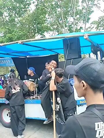 This is a video taken from a slightly elevated perspective, showing an outdoor scene. In the foreground, several young girls with their backs to the camera are visible. Behind them, a crowd of people, mostly adults and teenagers, are gathered. Many are wearing black attire. Some are looking towards the center of the scene, while others appear to be participating. A truck with a blue tarp covering its top is parked on the right side of the frame. This truck has a large speaker mounted on it and appears to be a stage or performance area. Several people are standing on and around the truck. One man on the truck is holding a microphone. Another man on the truck is holding a long stick. In the middle of the crowd, a group of individuals in traditional clothing are performing. One man in the center is playing a wind instrument. The environment appears to be a rural or semi-rural area with trees and vegetation in the background. The ground is uneven and appears to be made of dirt and gravel. The overall impression is of a community event or celebration.