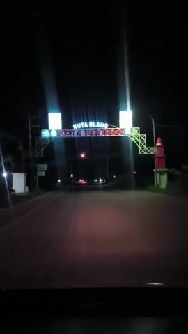 A nighttime view from inside a vehicle shows a road with streetlights and a prominent archway with illuminated text reading KUTA BLANG KOTA BERIMAN. The archway has decorative elements and possibly flags or signs on either side. A red and white lighthouse-like structure is visible to the right of the archway. The road ahead is dark, with car headlights creating streaks of light. The overall environment appears to be a town or city entrance.