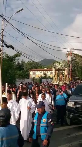 A procession is moving down a street lined with houses and utility poles. A large group of people are walking, many wearing white robes. In the center of the procession, a decorated float with figures is being carried. Several individuals in blue uniforms, appearing to be law enforcement or security, are also present, flanking the procession and walking alongside it. A black SUV is positioned on the right side of the frame, following the procession. The sky is partly cloudy, and a mountain is visible in the background.