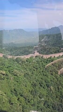 An aerial drone shot captures a vast, lush green mountainous landscape. The Great Wall of China, a long stone structure, snakes along the ridge of a hill, dotted with watchtowers. The wall is surrounded by dense forests, and distant mountains fade into a hazy blue sky. The scene is serene and majestic, showcasing the historical monument in its natural splendor.