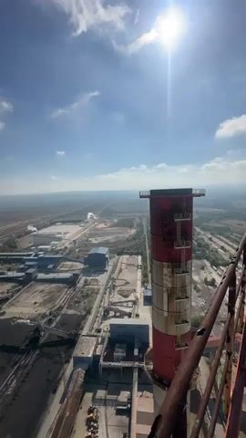 This is a vertical video taken from a high vantage point, likely an industrial structure such as a smokestack or tower. The foreground is dominated by the upper portion of a red and white striped industrial smokestack, with rusty metal structures visible on the right edge of the frame. Below, a sprawling industrial complex unfolds under a bright, sunny sky with scattered clouds. The complex includes various buildings, some with blue roofs, and large piles of dark material, possibly coal or mining waste, arranged in long rows. Conveyor belts and other industrial infrastructure are also visible. In the distance, the landscape stretches out, with more industrial activity and what appears to be a highway or railway line. The overall impression is one of a large, active industrial site on a clear day.