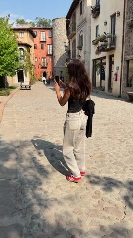 A woman with long brown hair is standing on a cobblestone street in a Mediterranean-style village, taking a picture with her phone. She is wearing a black tank top, grey jeans, and red sneakers. She has a black bag hanging off her shoulder. The street is lined with buildings that have stucco walls and tiled roofs. In the background, a golf cart-like vehicle is visible. The camera then pans to a nearby lawn which features three metal sculptures of donkeys, each fitted with saddles and carrying buckets. The donkeys are arranged in a row on a brick-laid path, and the lawn is surrounded by lush greenery.