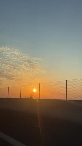 The video shows a sunset behind a chain-link fence, viewed from what appears to be a moving vehicle. The sun is low in the sky, glowing orange, and there are some patchy clouds above it. A grey concrete barrier is in the foreground and the road on which the vehicle is driving is visible at the bottom. Some utility structures and a distance marker can be seen sporadically through the fence as the vehicle progresses.