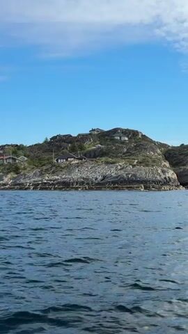 This media file captures a serene view of Hellesøy, Norway, from the water. The foreground is dominated by the deep blue, slightly rippled surface of the ocean, reflecting the bright daylight. In the midground, a rugged, rocky coastline rises, dotted with numerous houses, many featuring traditional Scandinavian architecture with dark roofs and light-colored walls. These homes are nestled into the sloping terrain, interspersed with patches of green vegetation and trees. The rocky outcrops suggest a natural, untamed landscape.

The background features a clear, pale blue sky with a few wispy white clouds, indicating fair weather. The overall scene is peaceful and picturesque, conveying a sense of tranquility and the beauty of a coastal Norwegian setting. The houses suggest a residential area, likely a small village or scattered dwellings along the coast. The time of day appears to be daytime, possibly late morning or early afternoon, given the bright, natural light. There is no visible text within the media file. The emotion evoked is one of calm and appreciation for natural and human-made harmony in a beautiful location.