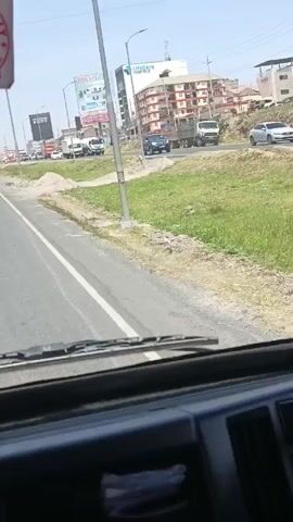 The video shows a view from inside a truck driving on a paved road in what appears to be a semi-urban environment. Several other vehicles, including cars, trucks, and a motorcycle, are visible on the road. The landscape adjacent to the road consists of some vegetation and piles of sand or gravel. A billboard advertising "Hikvision" and "LifeCare Hospitals" stands prominently in the background, next to several medium-rise buildings. The weather appears to be clear and sunny. The camera is fixed and captures the scenery from the driver's perspective through the truck's windshield.