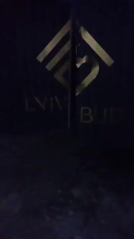 The video shows a dark blue surface with a gold logo and the words "LVIV BUD" written beneath. The logo is a stylized image consisting of geometric shapes and lines, resembling a building or architectural design. The surface appears to be part of a wall or structure, but the lighting is dim, making it difficult to discern specific details about the surrounding environment. The camera remains stationary throughout the video.