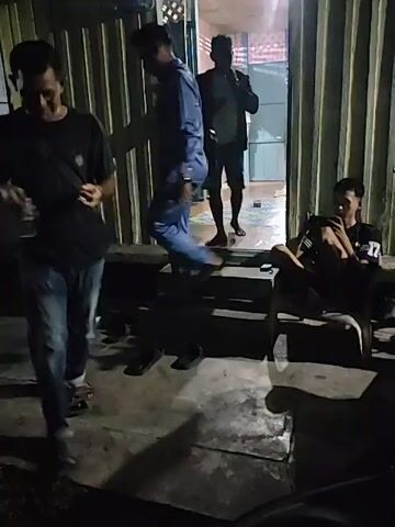 The video shows a dimly lit outdoor scene with several people. In the foreground, a person in a dark shirt and blue jeans walks towards the camera, their face obscured by motion blur. To their left, a broom leans against a corrugated metal wall. In the background, three men stand near an open doorway. One man in a blue suit walks down a set of stairs, and another man in a grey shirt and shorts stands talking to someone inside the doorway. A third man, partially visible, stands behind them. To the right, a young man sits on a bench, looking down at a phone. Several pairs of shoes are scattered on the ground. The environment appears to be a sidewalk or patio area in front of a building.
