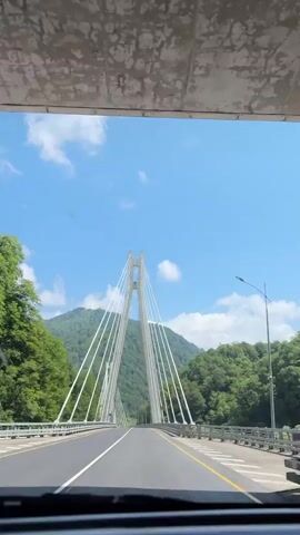 The video shows a first-person view from a vehicle driving on a road. In the foreground, the dashboard of the car is visible. The road ahead is paved and has lane markings. A modern cable-stayed bridge with tall white pylons and numerous cables dominates the center of the frame. The bridge spans across a valley. On either side of the road and bridge, lush green trees cover rolling hills and mountains. The sky above is a clear blue with scattered white clouds, suggesting a sunny day. The camera appears to be mounted on the dashboard or windshield of the vehicle, providing a straight-on perspective as the car approaches and drives onto the bridge. There are no other vehicles or people visible in the immediate vicinity on the road. The overall scene conveys a sense of travel through a scenic, mountainous landscape.