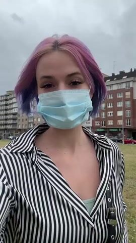 A young woman with a light blue disposable face mask and striking pink and purple hair is featured in a close-up, self-recorded video. She wears a black and white striped long-sleeved shirt. Initially, she looks directly at the camera, then turns her head to gaze to her right, and then returns her attention to the camera. Her hair, which is pink at the roots and purple towards the ends, is gently ruffled by the wind.

The setting is an open grassy area, possibly a park or field, in what appears to be an urban or suburban environment. In the background, multiple multi-story apartment buildings are visible under an overcast sky, suggesting a cloudy day. A street with parked cars can also be seen behind the buildings. The video captures a brief moment in Gijón, Spain. No visible text is present in the video.