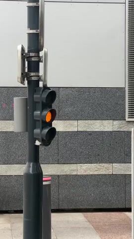 A traffic light pole stands on a sidewalk in front of a building. The traffic light, which consists of three black circular lenses stacked vertically, is dark. A gray cylindrical object is attached to the pole below the traffic light. Two reflective rectangular signs are mounted on the pole above the traffic light. The building facade behind the pole has alternating bands of light gray and dark gray speckled material.