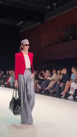 The video shows a fashion show with a model walking down a runway. The model is wearing a red blazer, a white top, gray wide-leg pants, a gray hat, and large sunglasses. She is also carrying a black tote bag and wearing pearl jewelry. The runway is white, and the audience is seated on either side, watching the model. The background shows a dimly lit theater with tiered seating. The model is walking with her head held high and a slight smile. The video captures the model from various angles as she walks.