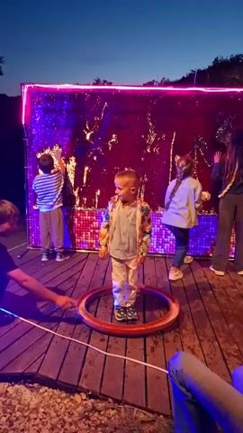 The video shows a child standing inside a large bubble on a wooden stage with others around. The stage is decorated with a sparkling backdrop lit by neon lights. A man is creating the bubble around the child. Other children stand near the backdrop, interacting with it. The sky is a dusky blue, suggesting it is evening.