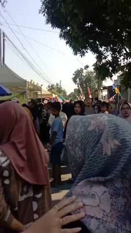 The video shows a crowded street scene, possibly a market or festival, with many people walking in both directions. Several tents or stalls are set up along the side of the street, and there are decorations like flags or banners visible in the background. The people are of various ages, including adults and children, and many are wearing casual clothing, with some women wearing headscarves. The lighting suggests it is daytime, with sunlight filtering through trees on the right side of the frame. The camera appears to be moving with the crowd, capturing a dynamic and bustling atmosphere.