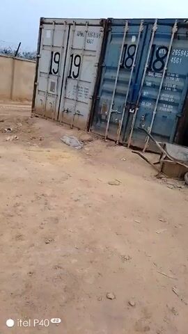 The video shows an outdoor scene with several shipping containers lined up in a row. The ground is dirt and there are scattered items, including a wheelbarrow, a tire, pieces of metal, hoses, cans, and boxes. There are no people visible in the video. The camera pans across the scene, providing a wide view of the area. The overall impression is that this is a storage or work area.