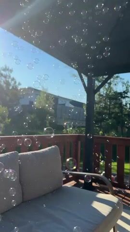 A deck with a sofa and railing is visible. Numerous bubbles float through the air, catching the sunlight. A house and trees are in the background under a clear blue sky.