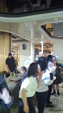 A group of people are dancing in a restaurant or party venue. Some are standing at tables, while others are actively dancing to upbeat music. The room has a reflective ceiling and visible lighting fixtures. There's a glimpse of a bar area with a mirror, and the overall atmosphere is festive. The people are dressed in casual attire, suggesting a relaxed social gathering.