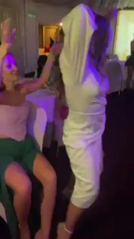 The video shows a person with long, dark, curly hair, wearing a white dress and white heels, dancing in a dimly lit room with a party atmosphere. The camera focuses on the person’s back as they move, occasionally showing other people in the background, including a person in a red dress and another in a black outfit with a wide-brimmed hat. Tables with white tablecloths are visible in the background, along with what appears to be decorative lights and white balloons on the ceiling. The lighting in the room changes from green to blue to purple, creating a club-like ambiance. The person in the white dress moves around the room, swaying and squatting low to the ground while dancing.